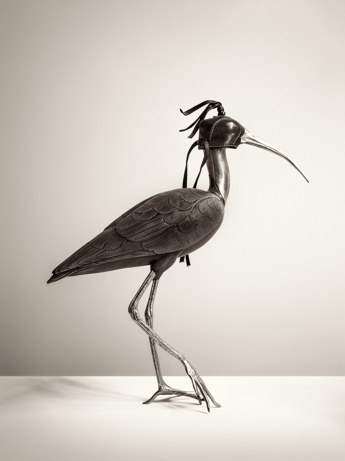 Escultura de un ave zancuda de pico largo y curvo, en metal oscuro, con una caperuza de halconeria de cuero cubriendole los ojos