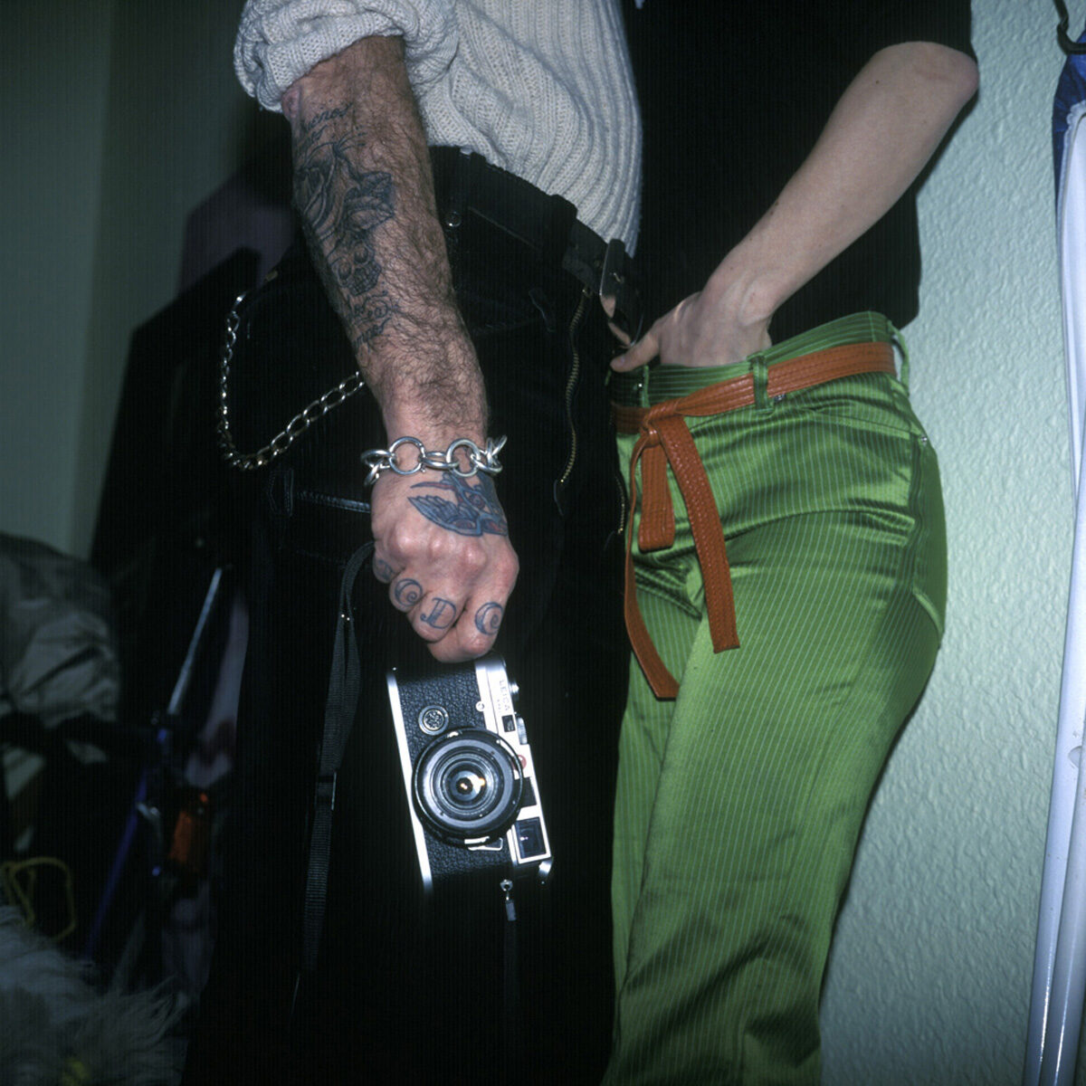 Dos personas de pie muy juntas, de cintura para abajo: una sostiene una cámara telemétrica y lleva tatuajes en la mano y el antebrazo; la otra viste un pantalón verde con cinturón naranja.