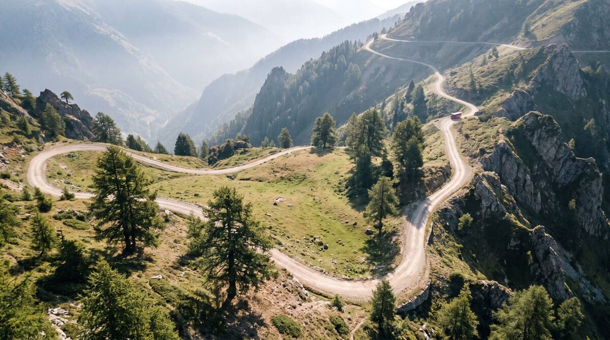 Vista aérea de una carretera de montaña con curvas cerradas entre bosque, con un autobús rojo pequeño al fondo.