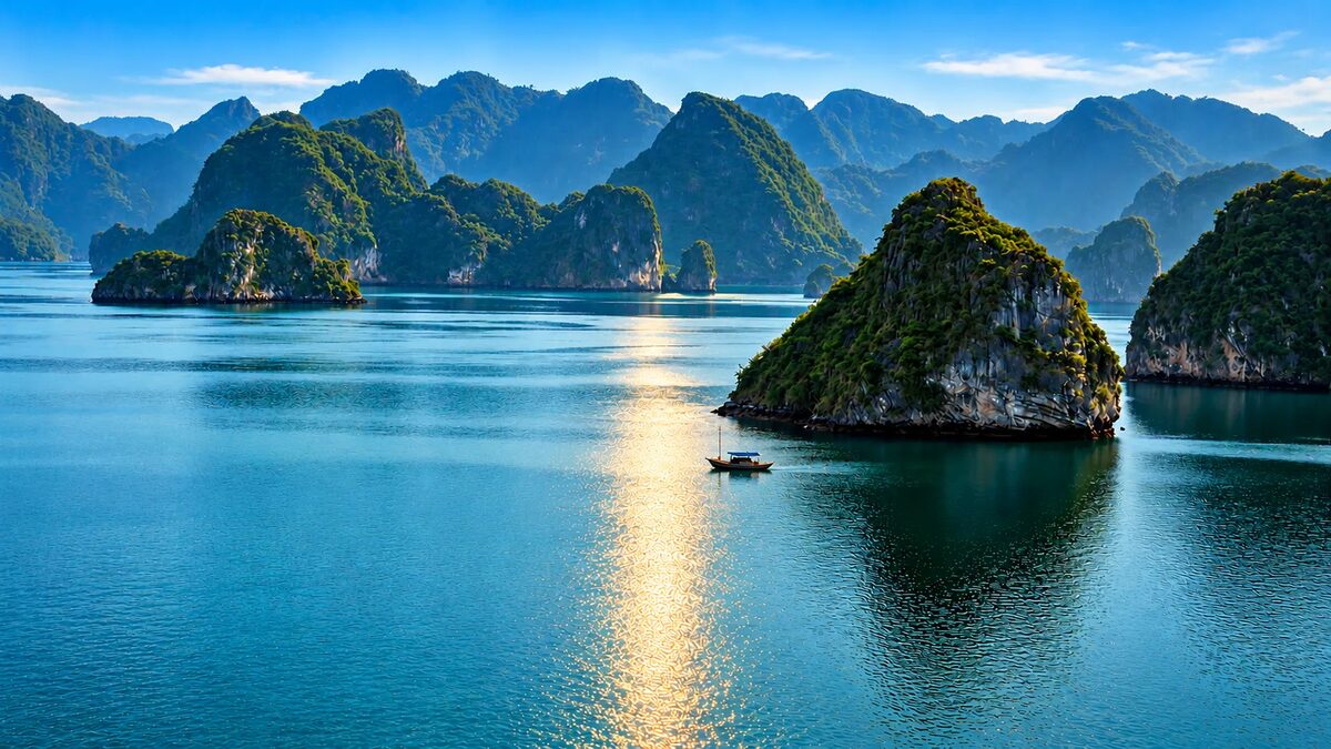 La bahía de Ha-Long con sus islas de roca cubiertas de vegetación, el agua azul y una barca pequeña con el reflejo del sol.