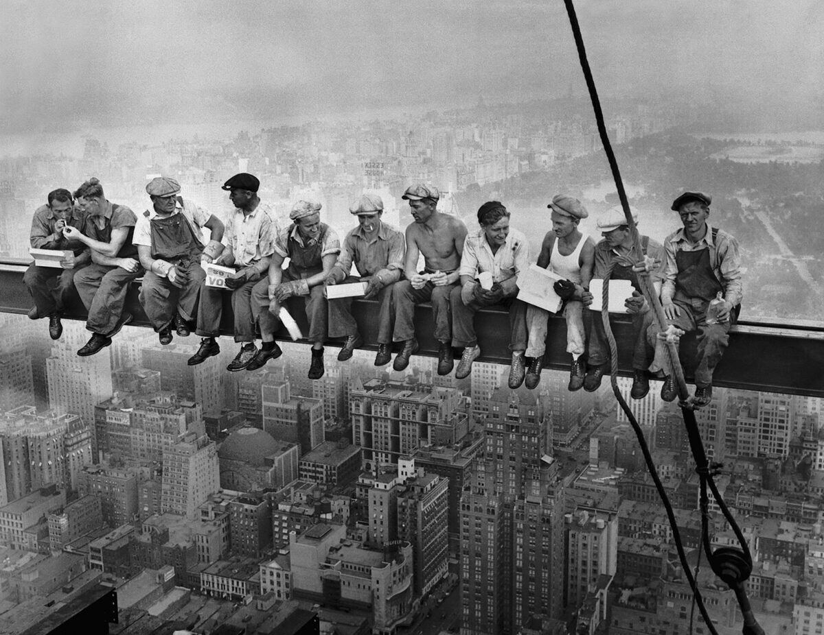 Once obreros almorzando sentados en fila sobre una viga de acero, con la ciudad muy por debajo.