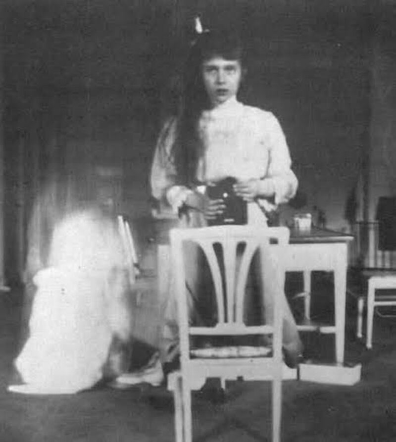 Una adolescente de principios del siglo XX sostiene una cámara de cajón a la altura del pecho y se retrata ante un espejo de marco oscuro.