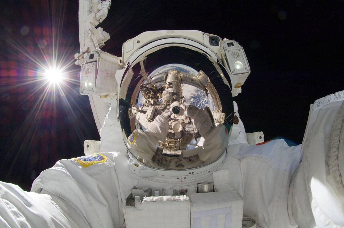 Un astronauta con traje blanco flota en el espacio; en el visor dorado de su casco se reflejan el Sol, la Tierra y el brazo robótico de la estación.
