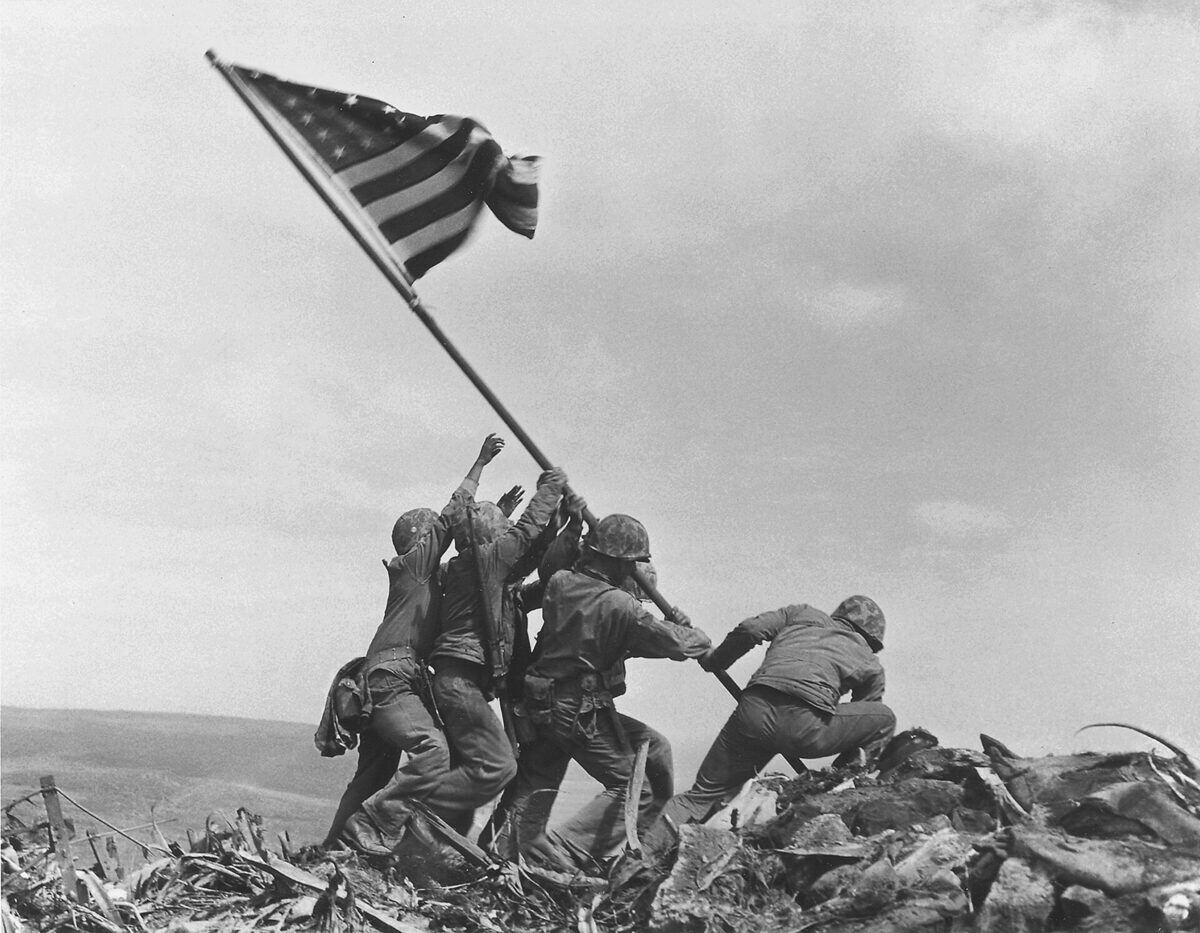 Seis marines empujando juntos el mástil de una bandera sobre un terreno de piedras.