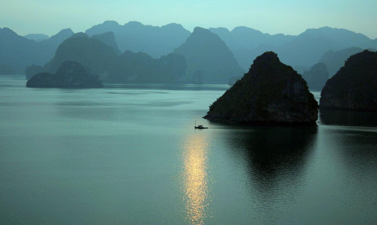 La bahía de Halong al atardecer, con el sol reflejado en el agua en calma y una barca solitaria.