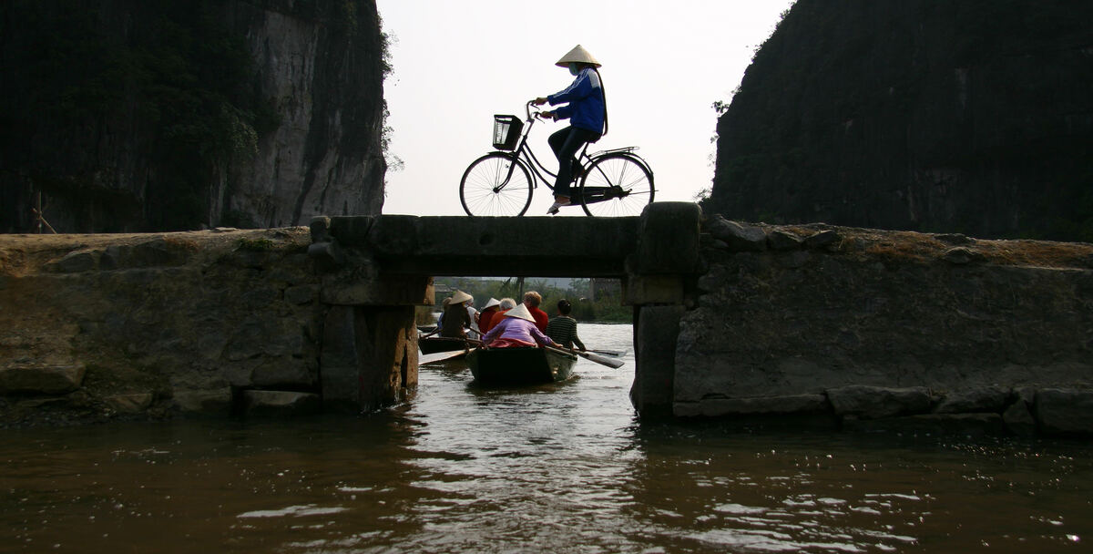 Un ciclista con sombrero cónico cruzando un puente de piedra sobre el agua, en Vietnam.
