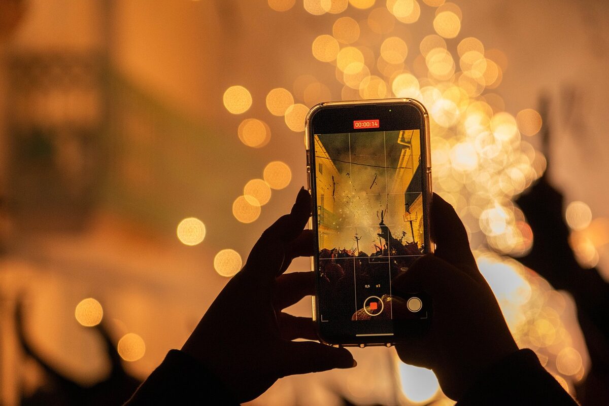 Unas manos sostienen un teléfono móvil que graba una celebración nocturna; en la pantalla se ve la escena ya procesada, entre luces desenfocadas.